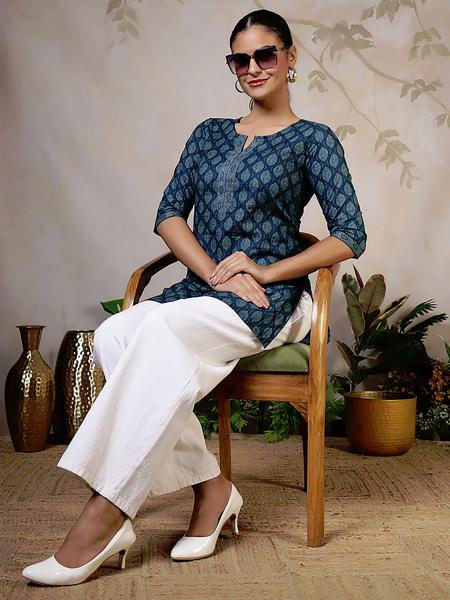 The model is wearing a petrol-colored tunic printed with a leaf pattern, combined with wide-legged, summery white trousers.